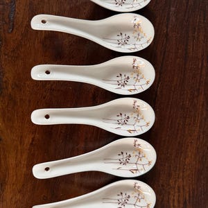 Könnte beinhalten: Sechs weiße Keramik-Suppenlöffel mit einem floralen Design in Braun und Gold. Jeder Löffel hat ein kleines Loch am Ende des Griffs. Die Löffel sind vertikal auf einer dunklen Holzoberfläche angeordnet.