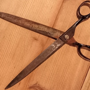 May include: A pair of vintage metal scissors with long, pointed blades showing signs of oxidation and wear. The scissors feature two black finger loops and a central screw mechanism connecting the blades, which are positioned in an open, crossed configuration.