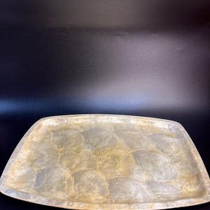 Peut inclure: Plateau décoratif rectangulaire avec une finition nacrée. Le plateau a des coins arrondis et une surface subtile et chatoyante avec un mélange de tons or et blanc. Le plateau est posé sur un fond sombre.