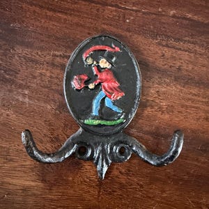 May include: Black cast iron wall hook with a decorative oval emblem featuring a colorful illustration of a person in a red coat and hat. The hook has two curved arms and a decorative base.