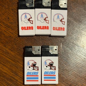 May include: A collection of seven white lighters featuring the Houston Oilers football team logo. Each lighter displays a helmet graphic with the word "OILERS" in blue. The lighters are arranged on a wooden surface.