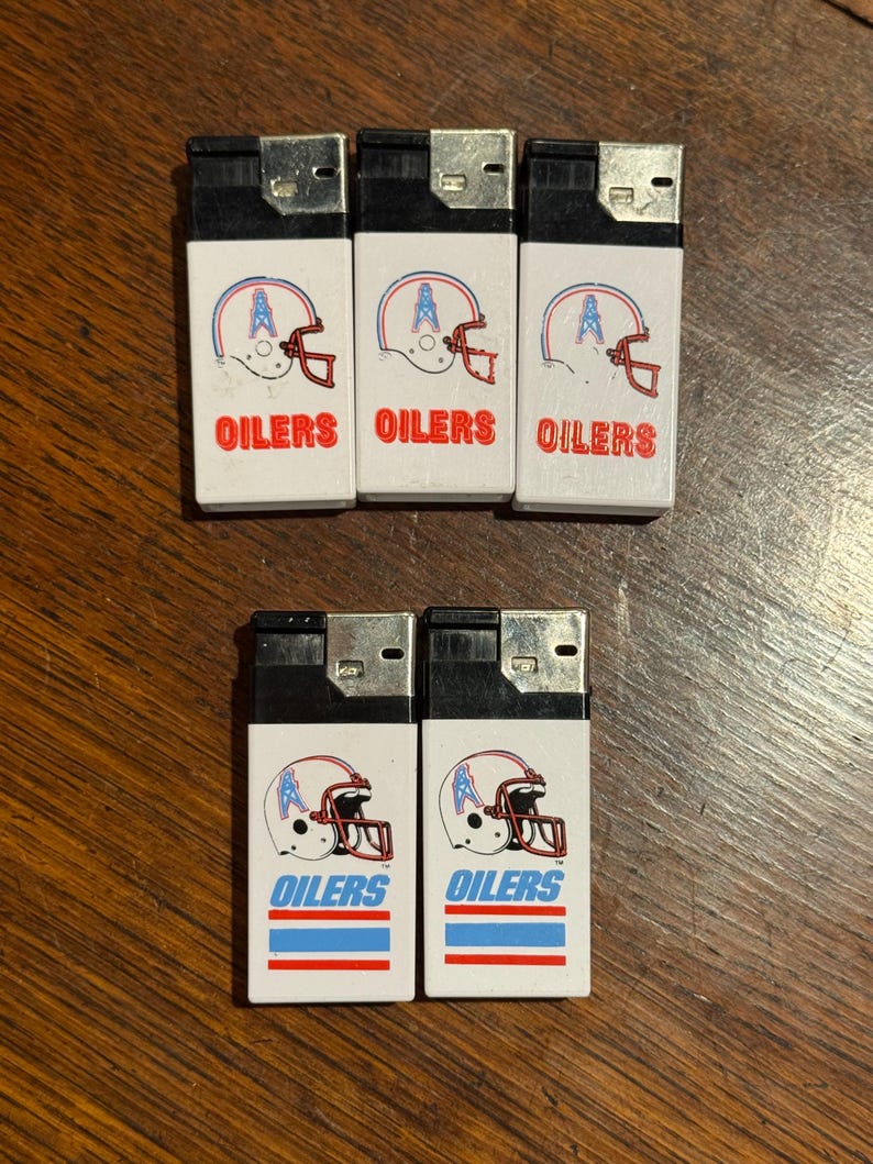 May include: A collection of seven white lighters featuring the Houston Oilers football team logo. Each lighter displays a helmet graphic with the word "OILERS" in blue. The lighters are arranged on a wooden surface.