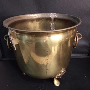 Pode incluir: Um vaso de latão polido com uma abertura larga e um corpo liso e arredondado. O recipiente possui duas alças laterais circulares e apoia-se em três pés curvos e ornamentados. A superfície metálica apresenta uma pátina natural.