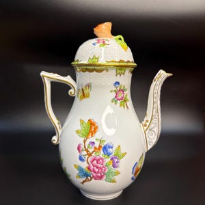 May include: White porcelain teapot with floral and butterfly designs. The teapot features a curved handle, a spout, and a lid with a flower-shaped knob. Gold trim accents the edges. The teapot is decorated with colorful flowers and butterflies.
