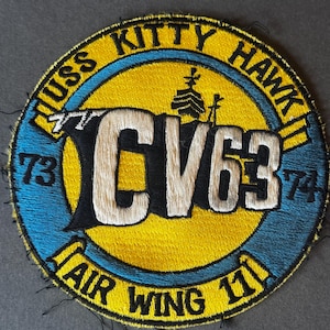 May include: Embroidered patch with the text "USS KITTY HAWK CV63 AIR WING 11". The patch is circular, with a yellow center and blue and yellow border. The text is in white and black.