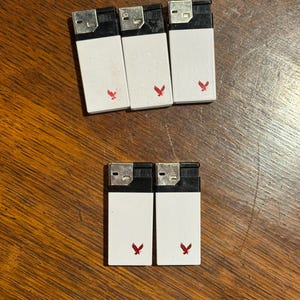 May include: A collection of seven white lighters with black tops and a red eagle logo. The lighters are arranged on a wooden surface. The lighters are rectangular and have a simple, functional design.