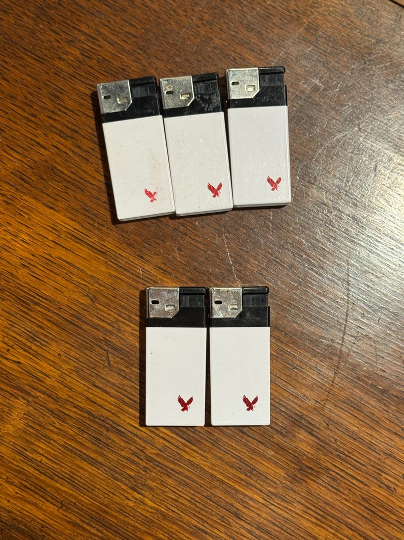 May include: A collection of seven white lighters with black tops and a red eagle logo. The lighters are arranged on a wooden surface. The lighters are rectangular and have a simple, functional design.
