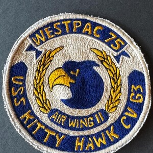 May include: Embroidered patch with a circular design. The patch features text that reads "WESTPAC 75", "USS KITTY HAWK CV 63", and "AIR WING II". The central design includes a blue eagle head and gold wheat stalks.