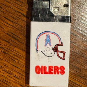 May include: A vintage white lighter featuring the Houston Oilers logo. The logo includes a football helmet with a blue and red outline, an oil derrick, and the word "OILERS" in red. The top is black and silver.