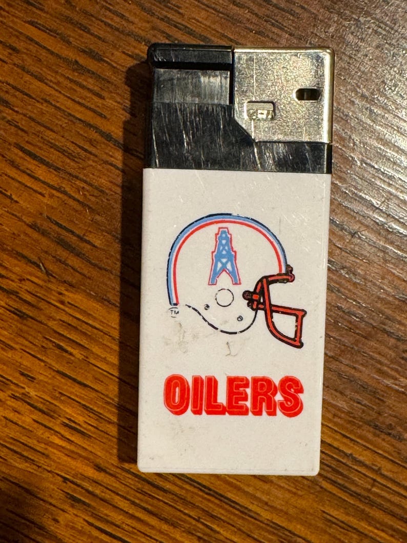 May include: A vintage white lighter featuring the Houston Oilers logo. The logo includes a football helmet with a blue and red outline, an oil derrick, and the word "OILERS" in red. The top is black and silver.