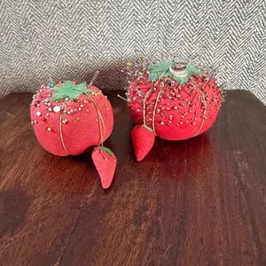 Puede incluir: Dos cojines rojos con forma de tomate con detalles de hojas verdes y formas de fresa de tela adjuntas. Los cojines están cubiertos de alfileres y están sobre una superficie de madera oscura.