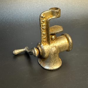 Op de afbeelding: Een antieke, gietijzeren vleesmolen met een klemontwerp. De molen heeft een aangetaste, goudbruine afwerking, een handslinger en een cilindrische maalruimte. De klem is ontworpen om aan een tafel- of aanrechtblad te bevestigen.