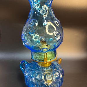 Könnte beinhalten: Eine blaue Vintage-Öllampe aus Glas mit einem dekorativen Blumenmuster. Die Lampe hat einen runden Sockel, einen bauchigen Körper und einen gerüschten Aufsatz. Sie hat einen Griff und Messingakzente. Die Lampe ist etwa 25 cm hoch.