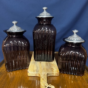Puede incluir: Tres tarros de vidrio morado oscuro con tapas plateadas. Los tarros tienen un diseño acanalado y están colocados sobre una tabla de cortar de madera y una mesa de madera. Las tapas tienen un patrón decorativo.