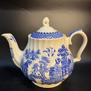 Puede incluir: Una tetera de cerámica blanca con un patrón azul de estilo willow que muestra pagodas, sauces y puentes. La tetera tiene un cuerpo acanalado, un asa curva, un pico vertedor y una tapa a juego con un remate en la parte superior.