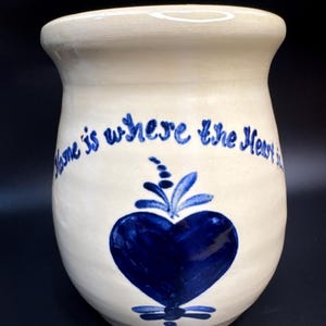 May include: Cream-colored ceramic vase with a blue heart design and the phrase "Home is where the Heart is" in blue script. The vase has a rounded body and a slightly flared rim.
