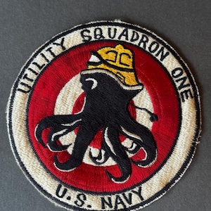 Könnte beinhalten: Gestickter Aufnäher mit einem schwarzen Oktopus, der einen gelben Hut trägt, auf einem rot-weißen Zielscheibenmuster. Der Text "UTILITY SQUADRON ONE" und "U.S. NAVY" ist kreisförmig um den Oktopus angeordnet.