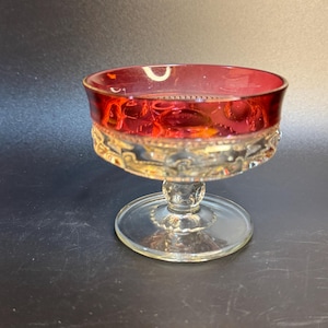 May include: A vintage, stemmed glass with a ruby red rim and a clear, ornate base. The bowl of the glass is a deep red, transitioning to clear glass with decorative detailing. The foot is a clear, circular base.