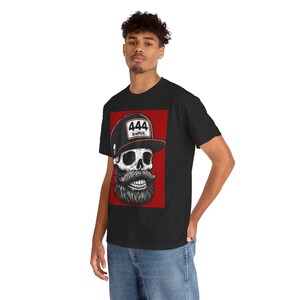 May include: A black t-shirt featuring a graphic print of a skull with a thick beard wearing a black baseball cap with the text 444 and GraffitiX on the front. The graphic is set against a rectangular red background on the chest of the shirt.