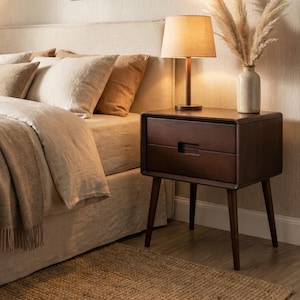 Puede incluir: Una mesita de noche de madera de color marrón oscuro con dos cajones y patas cónicas. La mesita presenta un marco rectangular con un diseño de tirador empotrado en los cajones. Está situada junto a una cama con una lámpara y un florero.