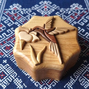 Könnte beinhalten: Hellbraune Holzbox mit einem geschnitzten Kolibri-Motiv. Der Kolibri, aus dunklerem Holz, ist im Flug dargestellt und trägt eine Blume. Die sechseckige Box steht auf einem blauen Stoff mit Muster. Maße in Zentimetern.
