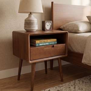 Puede incluir: Una mesita de noche de madera marrón con esquinas redondeadas, cuatro patas cónicas y un cajón. Encima hay una lámpara de color crema con base estampada y un reloj cuadrado de madera. Dentro hay dos libros titulados Dunes and Dust y Shapes in Space.