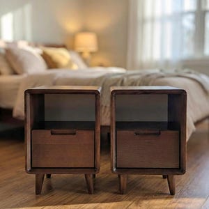 May include: Two matching wooden nightstands with a warm brown finish. Each nightstand has a drawer and an open shelf. The nightstands have rounded corners and tapered legs. The background shows a bed with pillows and a lamp.