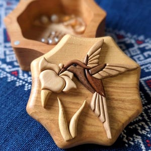 Könnte beinhalten: Holzschmuckschatulle mit einem Kolibri-Design. Der Deckel zeigt eine detaillierte Schnitzerei eines Kolibris im Flug, der sich von einer Blume ernährt. Die Schatulle ist aus hellem und dunklem Holz gefertigt und hat eine sechseckige Form.