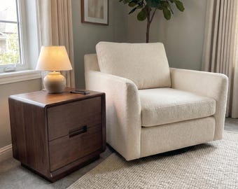 May include: A dark brown wooden nightstand with two drawers and a rounded edge design, topped with a small table lamp featuring a cream-colored shade and a rounded base, positioned next to a cream-colored upholstered armchair with a textured fabric.