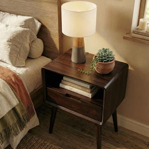 May include: A dark brown wooden nightstand with a single drawer and an open storage shelf. The nightstand features rounded corners and four tapered legs. A cylindrical table lamp with a white shade and a succulent in a terracotta pot sit on top.