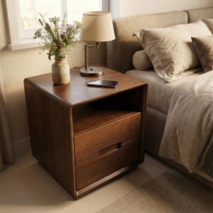May include: A dark brown wooden bedside table with a drawer and open shelf. A small lamp with a beige shade and a smartphone sit on top. A vase of flowers is also on the table. The table is next to a bed with beige bedding.