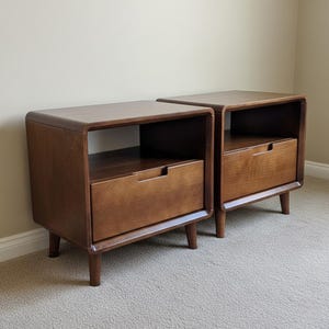 May include: A pair of wooden bedside tables in a mid-century modern style. Each table features a drawer with a recessed handle and an open shelf below. The tables are made of dark brown wood and have tapered legs. The design is sleek and functional.