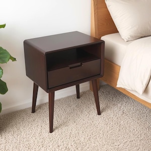 Puede incluir: Una mesita de noche de madera de color marrón oscuro con una superficie rectangular, un estante de almacenamiento abierto y un cajón extraíble con tirador empotrado. Se apoya sobre cuatro patas cónicas que se extienden hacia afuera.