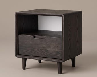 May include: A dark brown wooden bedside table featuring a square frame with rounded corners, an open storage compartment at the top, and a single pull-out drawer below. The piece is supported by four tapered legs and displays a visible wood grain.