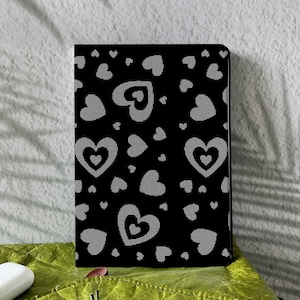 May include: A black notebook featuring a repeating pattern of white hearts. The hearts are of different sizes and are spread across the cover. The notebook is rectangular and rests on a green surface.