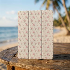 Può includere: Custodia per tablet con motivo floreale di rose rosa su sfondo crema. La custodia è realizzata in materiale resistente e ha un design a tre ante. È appoggiata su un tavolo di legno, con una spiaggia e l'oceano sullo sfondo.