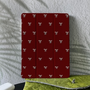 May include: A burgundy tablet case featuring a repeating pattern of small white bows. The case is closed and has a tri-fold design. The background is a neutral grey with a plant shadow.