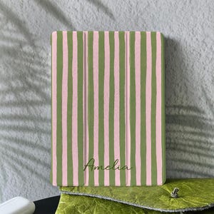 Può includere: Una custodia per tablet a righe verdi e rosa, con il nome "Amelia" in corsivo. Poggia su una pochette in pelle verde. Lo sfondo è un muro grigio chiaro con ombre.
