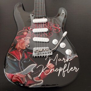 Könnte beinhalten: Eine schwarze E-Gitarre mit glänzendem Finish, mit einem Porträt von Mark Knopfler und dem Text "Mark Knopfler" in weißer Schrift. Die Gitarre hat weiße Tonabnehmer und eine silberne Brücke.