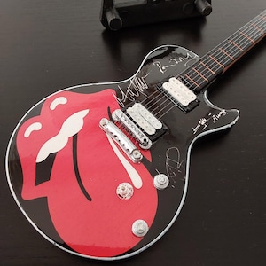 Könnte beinhalten: Miniatur-E-Gitarre in Schwarz mit dem roten Zungenlogo der Rolling Stones. Die Gitarre hat silberne Hardware, ein Griffbrett und Unterschriften. Ein kleiner schwarzer Ständer befindet sich im Hintergrund.
