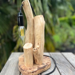 Peut inclure: Lampe de table en bois faite à la main, avec une ampoule, un cordon noir et un interrupteur. La lampe est fabriquée à partir de morceaux de bois naturels, dont une base et des supports verticaux. L'ampoule a un design de filament décoratif.