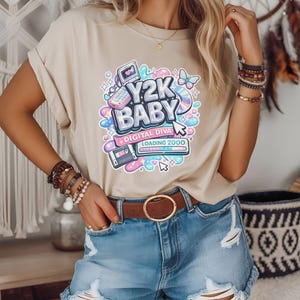May include: Beige t-shirt with a colorful graphic that reads "Y2K BABY DIGITAL DIVA LOADING 2000". The graphic includes a computer, calculator, cassette tape, and butterfly. The model is wearing denim shorts and a brown belt.