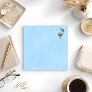 May include: A square notepad featuring a light blue sky and a watercolour hot air balloon in the upper right. The notepad is surrounded by stationery, including a gold watch, pen, and glasses.