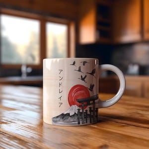 Op de afbeelding: Witte keramische mok met een Japans landschapsontwerp. Het ontwerp bevat een rode zon, een berg, een traditionele poort en vliegende vogels. De mok heeft een wit oor en de tekst "アンドレイア" aan de zijkant.
