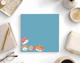 May include: A square, light blue notepad featuring four cartoon sushi illustrations. The sushi pieces include salmon, tuna, and a sushi roll. The notepad is surrounded by a gold watch, jewellery, a pen, glasses, and a cup of coffee.