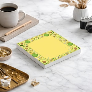 May include: A square notepad with a yellow center and a border of cartoon avocados and bees. The notepad is on a white surface with other office supplies, including a coffee mug, pen, and camera.