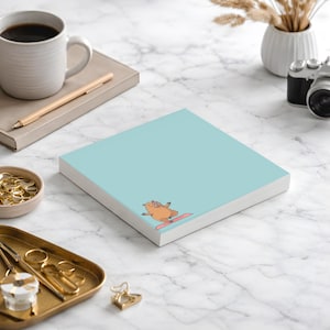 May include: A square, light blue notepad with a cartoon pig doing yoga on a pink mat. The notepad is on a white surface with a coffee mug, pen, and other office supplies.