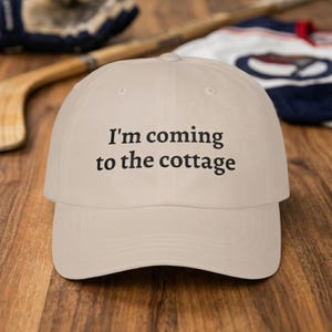 Peut inclure: Casquette de baseball beige avec le texte "I'm coming to the cottage" en noir. La casquette est faite d'une matière douce et a une visière incurvée. L'arrière-plan comprend une crosse de hockey, un gant de hockey et un maillot.