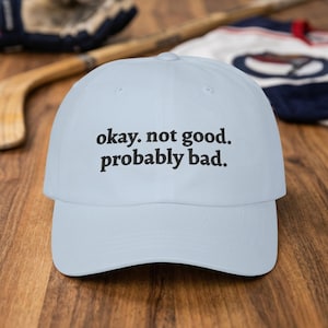 May include: A light blue baseball cap with the text "okay. not good. probably bad." in black. The cap has a curved brim and a classic six-panel design. The background includes a hockey stick, a hockey glove, and a jersey.