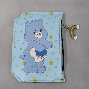 May include: A light blue wallet featuring a cartoon bear with a grumpy expression, surrounded by yellow stars. The wallet has a black zipper and a rainbow charm. The bear has a heart on its rear and a cloud with hearts on its belly.
